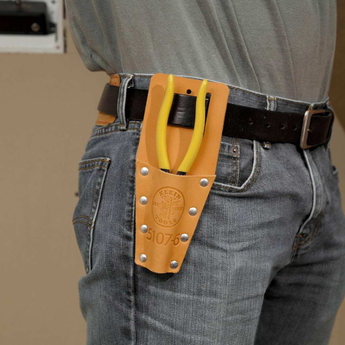 Lifestyle image showing a Klein Tools leather tool carrier attached to a professional's belt, demonstrating the convenient on-hip storage for pliers while working on-site.