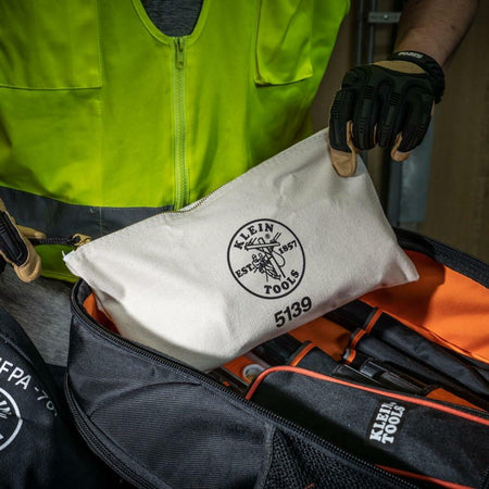 Person wearing safety gear holding Klein Tools canvas zipper pouch labeled "5139," placing it into a branded tool backpack in a jobsite setting.