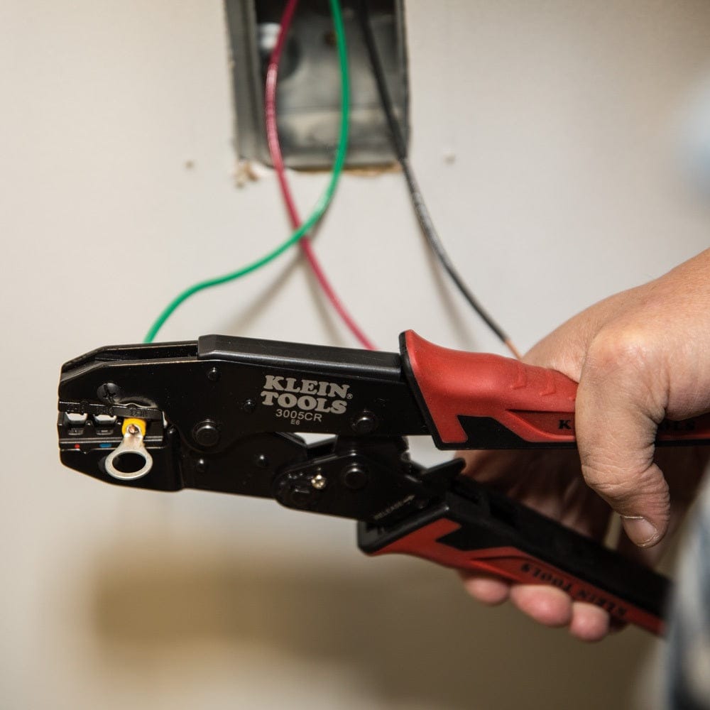 An action shot showing the crimping tool held near an electrical junction box with green and red wires, illustrating its use in residential or industrial wiring applications.