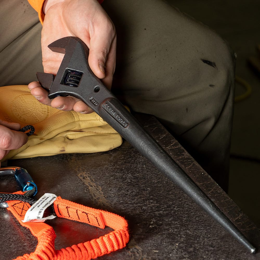 Worker holding a Klein Tools 3227 Adjustable Spud Wrench with tapered handle on a metal surface alongside gloves and safety gear, used for bolt alignment in steel construction.