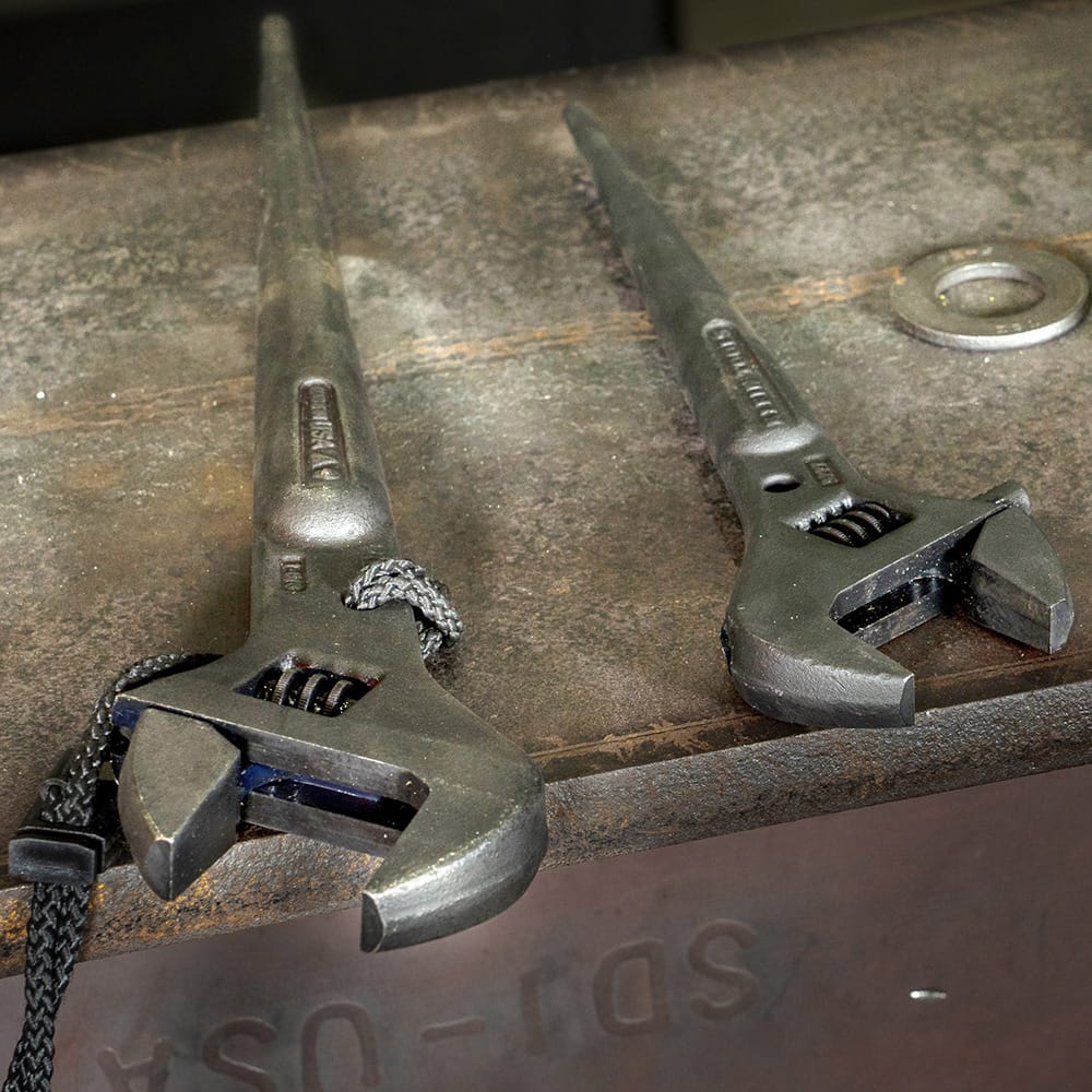 Two Klein Tools adjustable spud wrenches with long tapered handles on a metal workbench, one attached to a safety lanyard, used for bolt alignment and heavy‑duty construction fastening.
