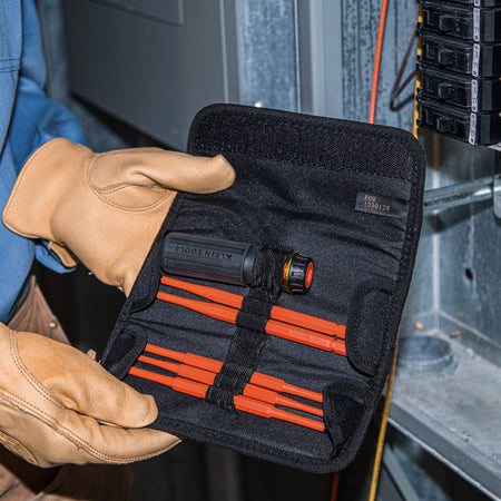 An electrician holding an open Klein Tools black canvas tool pouch displaying the complete 8-in-1 insulated interchangeable screwdriver set stored securely in elastic bands, demonstrating active field deployment of 1000-volt rated flame resistant drivers next to an open commercial breaker breaker box matrix.
