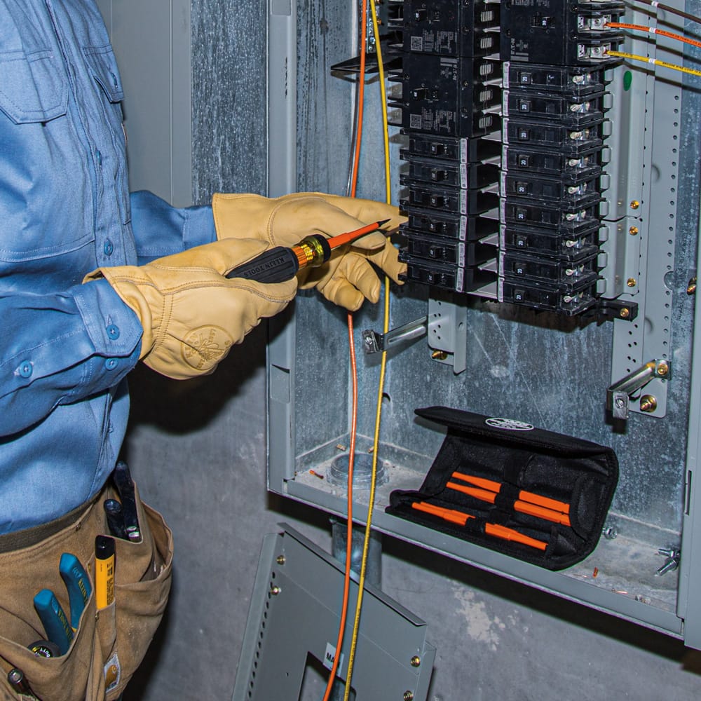 An electrician wearing tan leather safety gloves utilizes a fully assembled Klein Tools 1000V rated insulated interchangeable screwdriver to complete diagnostics inside a live circuit breaker panel matrix, featuring an open black canvas tool kit pouch with extra orange slim-tip replacement blades resting nearby.