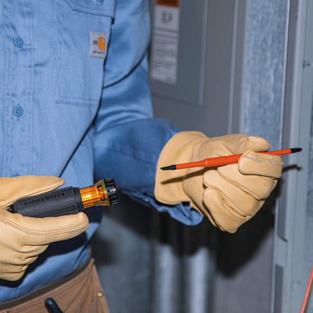 An electrician wearing heavy duty tan leather safety gloves holds a Klein Tools insulated screwdriver handle in one hand and an orange double-ended slim-tip blade in the other, demonstrating the quick-change twisting collar mechanism for swapping out bits while performing live panel maintenance diagnostics.