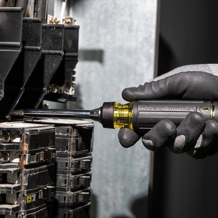 A gloved hand using the ratcheting screwdriver to tighten a screw on a circuit breaker inside an electrical panel.
