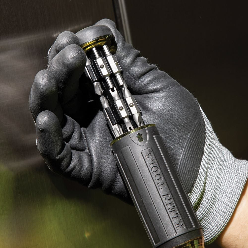 A close-up action shot showing a professional's gloved hand holding the Klein 32307 screwdriver with the bit storage compartment extended, ready for a specialty bit change.