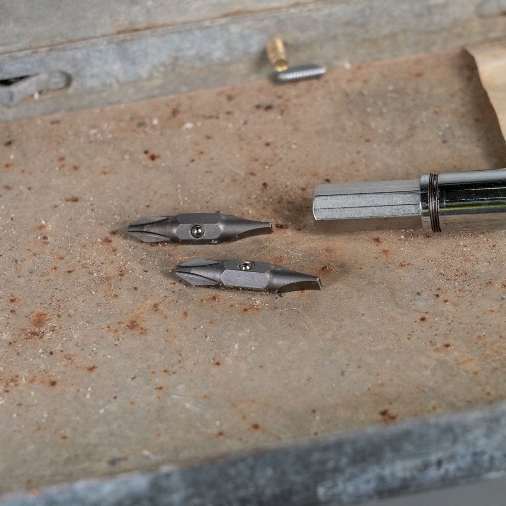Two replacement screwdriver bits resting on a weathered metal surface next to a chrome screwdriver shaft.