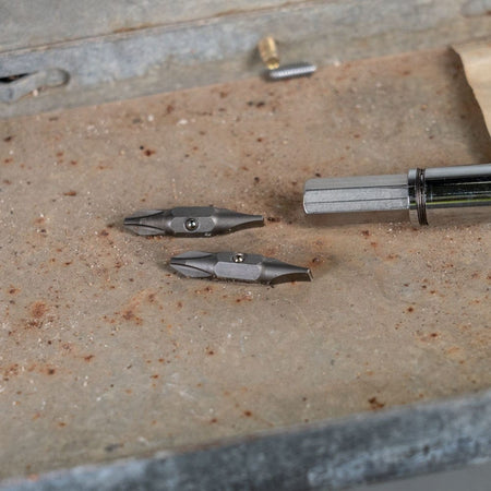 Two replacement screwdriver bits resting on a weathered metal surface next to a chrome screwdriver shaft.