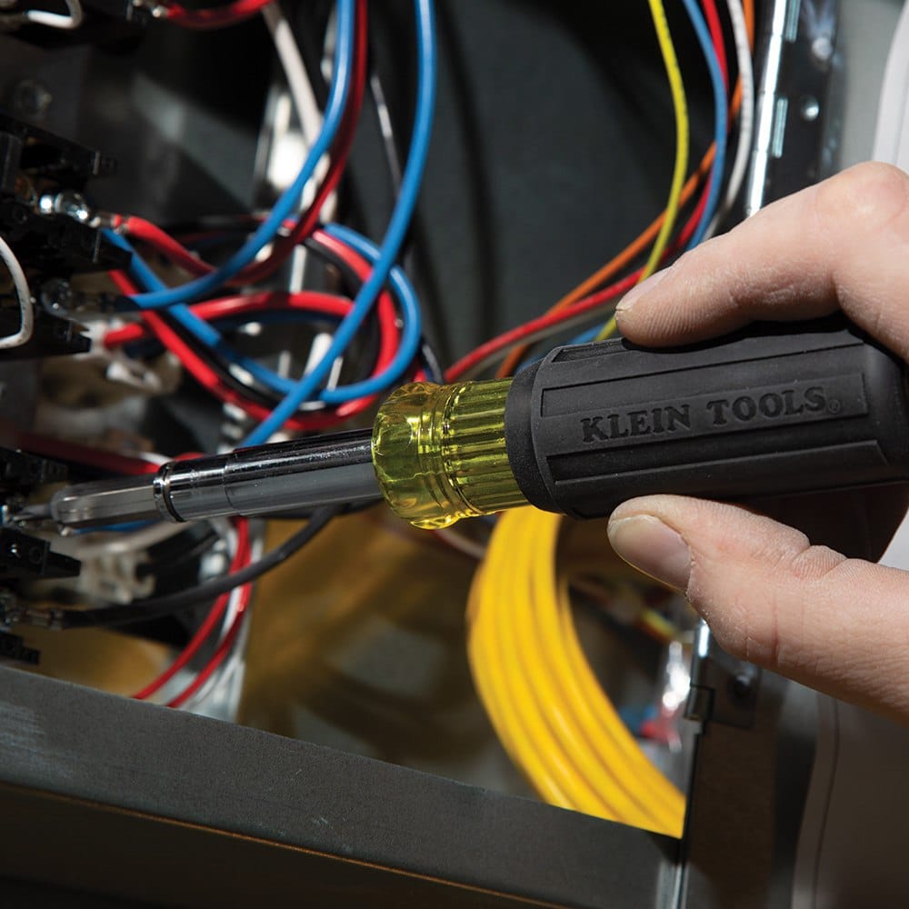 Close-up action shot of a professional using the 11-in-1 magnetic driver to tighten a terminal screw inside a complex electrical panel filled with multi-colored wiring.