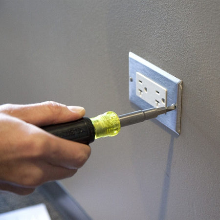 A person's hand using the Klein Tools multi-bit screwdriver to tighten a screw on a stainless steel electrical outlet wall plate.
