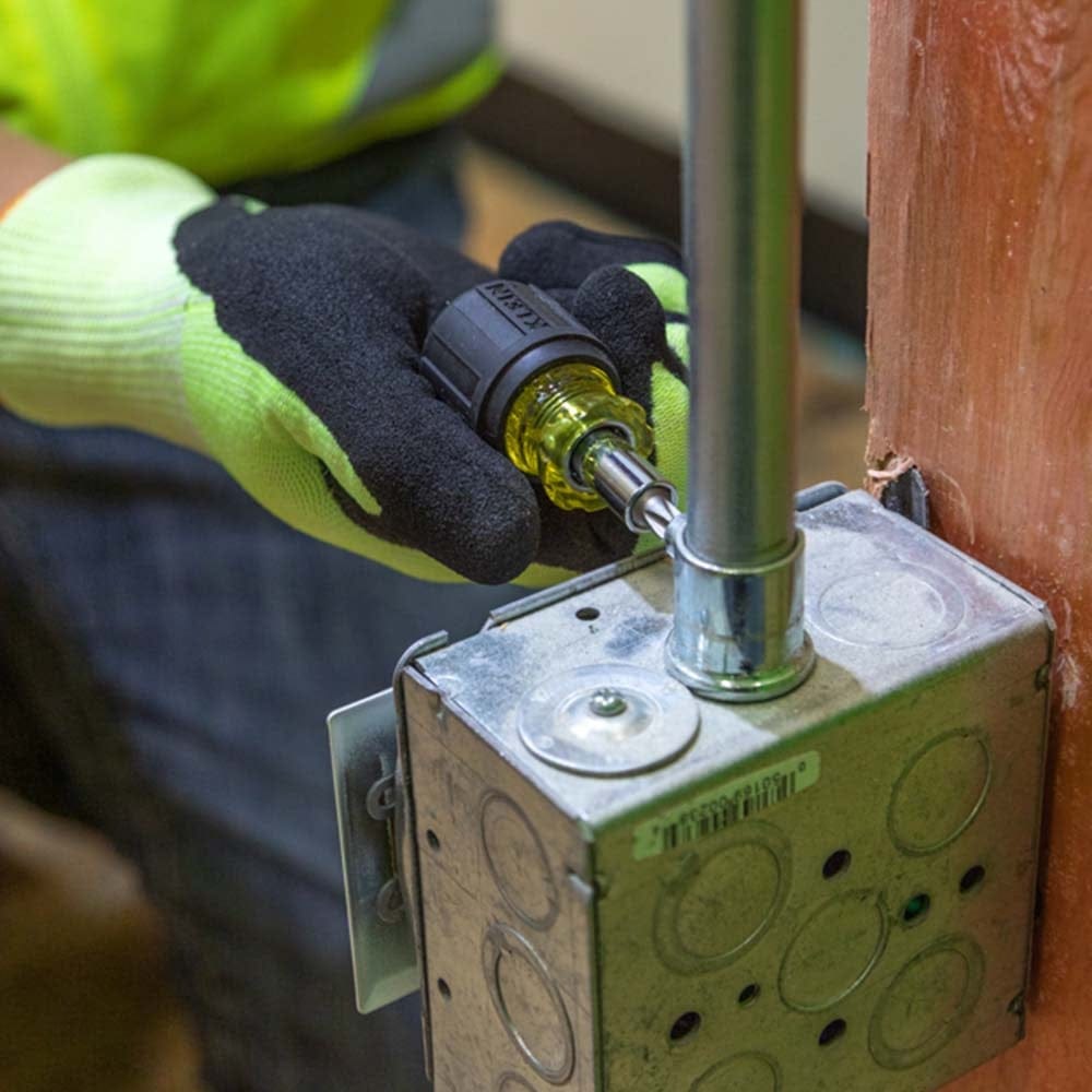 A worker wearing a high-visibility glove using the stubby screwdriver to tighten a conduit connector screw on a metal junction box.