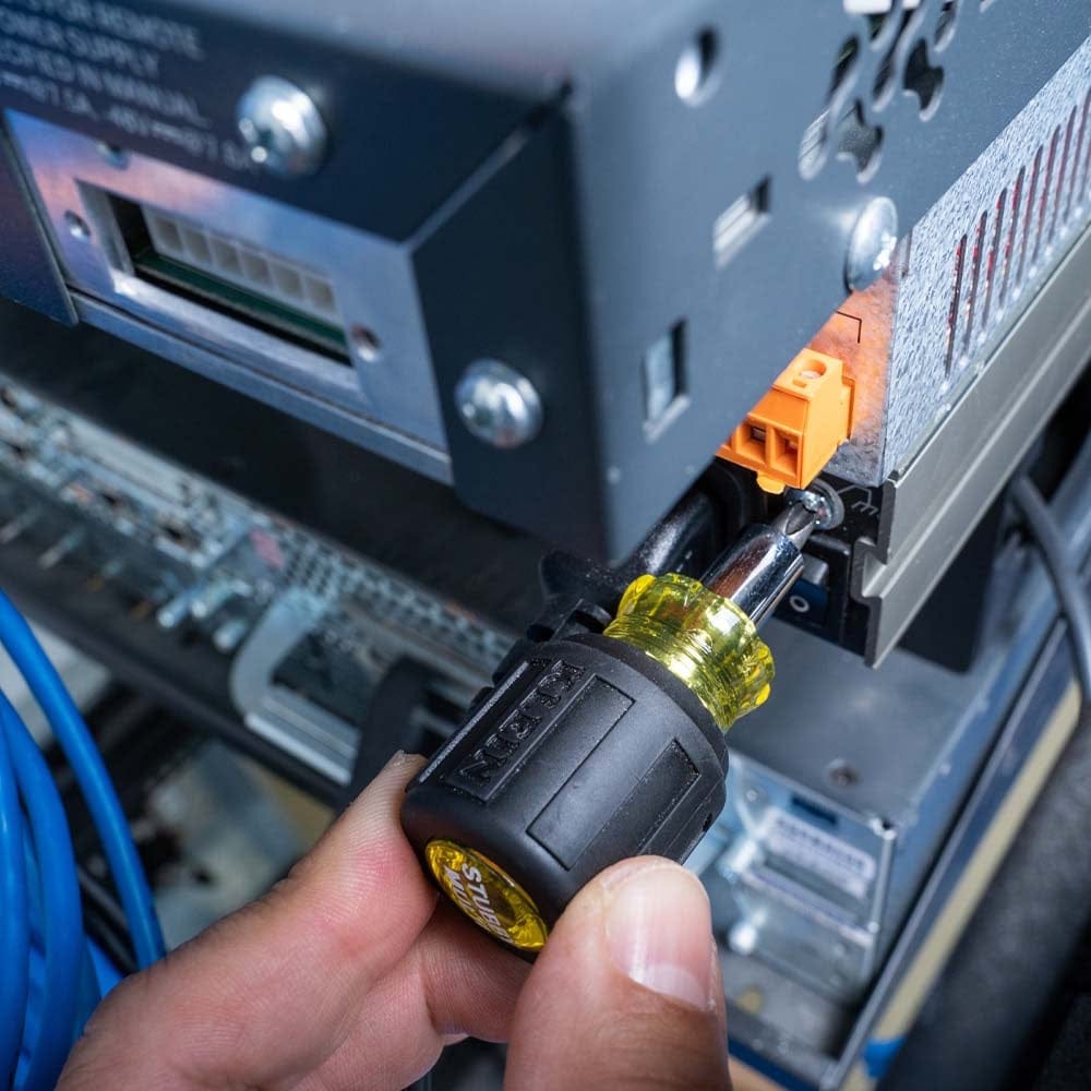 A close-up of a hand using the stubby screwdriver to secure a terminal connection on the back of a piece of rack-mounted electronic equipment.