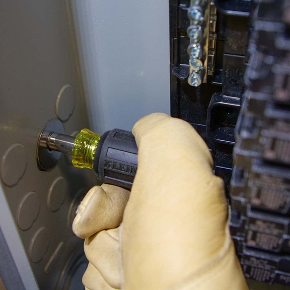 A gloved hand using the compact stubby screwdriver to tighten a terminal screw inside a grey electrical panel.