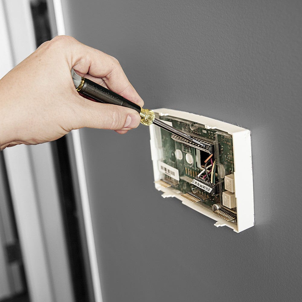 An action shot of a person using the 4-in-1 electronics screwdriver to make a fine adjustment on the internal circuitry of a wall-mounted digital thermostat.