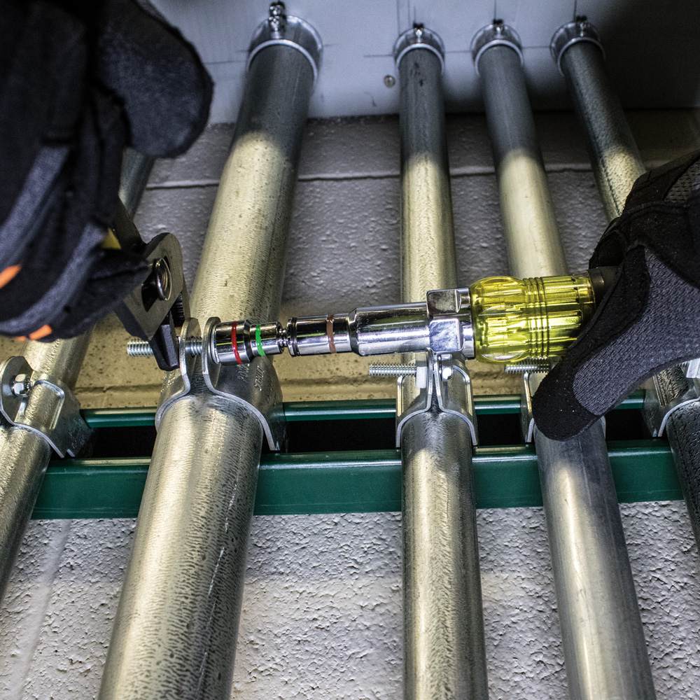 An action photograph showing the Klein 32807MAG magnetic nut driver being used with a pair of pliers for wrench-assist leverage to tighten a conduit clamp on a series of metal pipes.