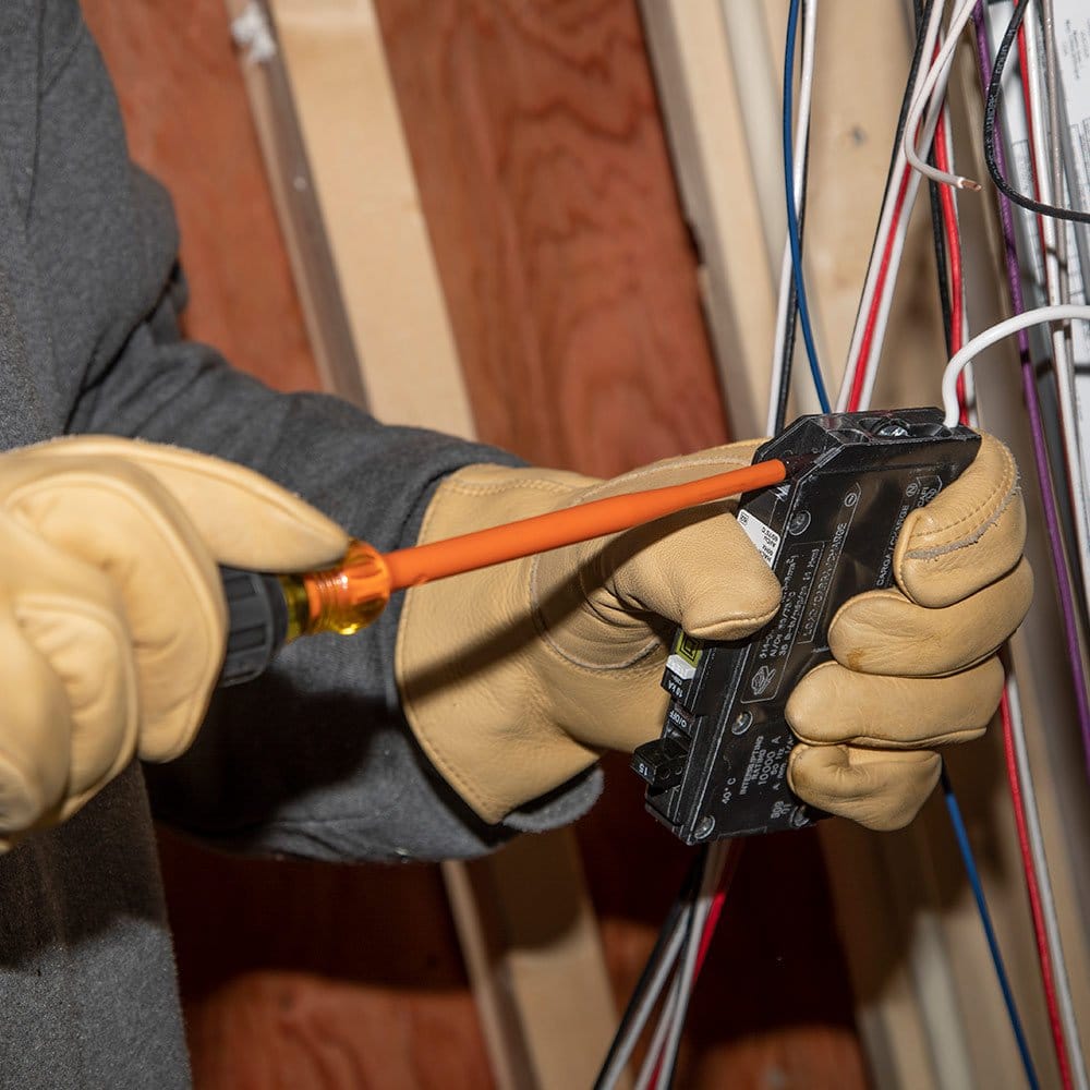 A tradesperson in gray work gear using a Klein 33734INS insulated tool to wire a slim-style circuit breaker, demonstrating the precision and safety of the 1000V rated slim-tip blade.
