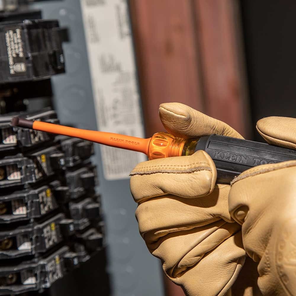 A close-up action shot of a professional wearing tan leather work gloves using an orange Klein slim-tip insulated screwdriver to adjust a circuit breaker in a crowded electrical box.