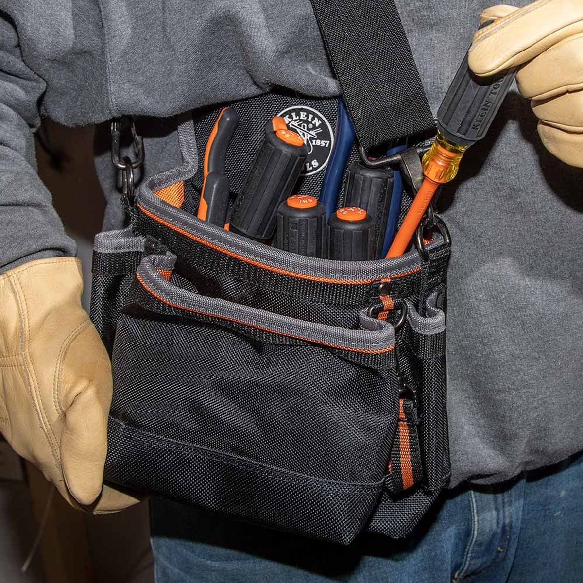 An electrician wearing durable tan leather safety work gloves pulls a 1000V rated orange slim-tip insulated screwdriver out of a heavy-duty black and orange canvas tool belt pouch packed with various high-dielectric hand tools, demonstrating real-world utility and jobsite organization.
