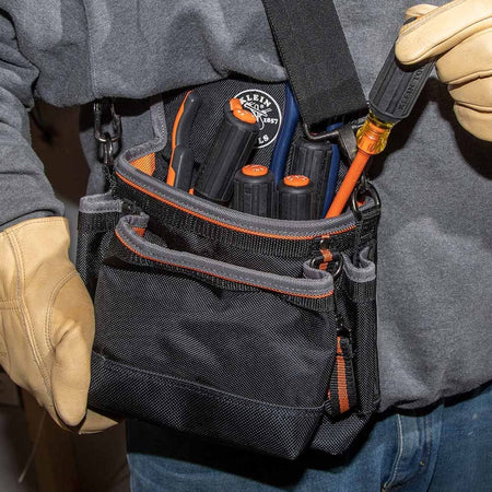 An electrician wearing durable tan leather safety work gloves pulls a 1000V rated orange slim-tip insulated screwdriver out of a heavy-duty black and orange canvas tool belt pouch packed with various high-dielectric hand tools, demonstrating real-world utility and jobsite organization.