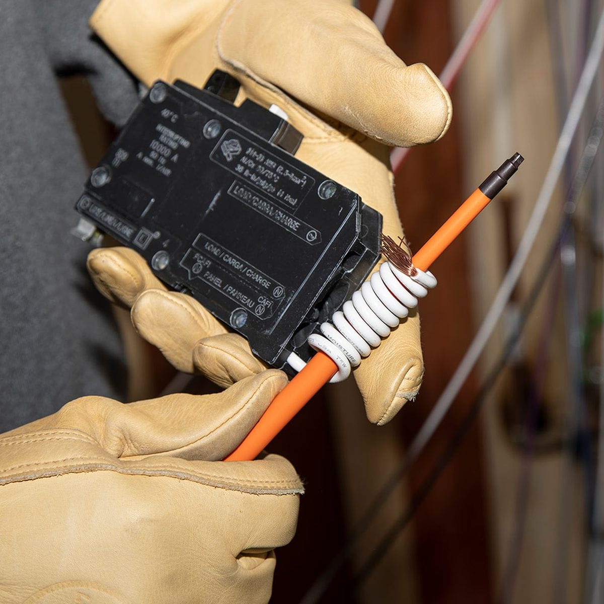 A close-up action view of a technician wearing protective tan leather gloves using a Klein Tools slim-tip insulated square-drive screwdriver to wire a commercial circuit breaker, demonstrating how the flush orange dielectric insulation safely passes into deep wire clamp ports without chipping.