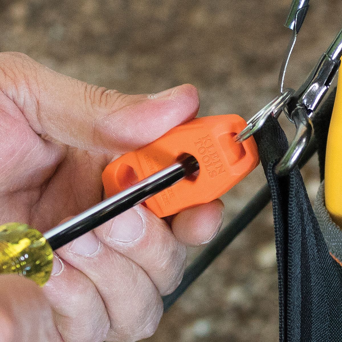 A close-up view of a trade professional passing a steel screwdriver shaft through the center hole of an orange Klein Tools magnetizer demagnetizer block accessory attached to a tool pouch by a metal key ring, illustrating the immediate magnetic charging process on a jobsite.