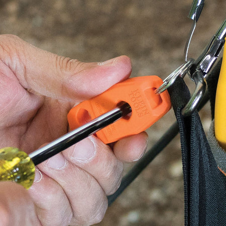 A close-up view of a trade professional passing a steel screwdriver shaft through the center hole of an orange Klein Tools magnetizer demagnetizer block accessory attached to a tool pouch by a metal key ring, illustrating the immediate magnetic charging process on a jobsite.