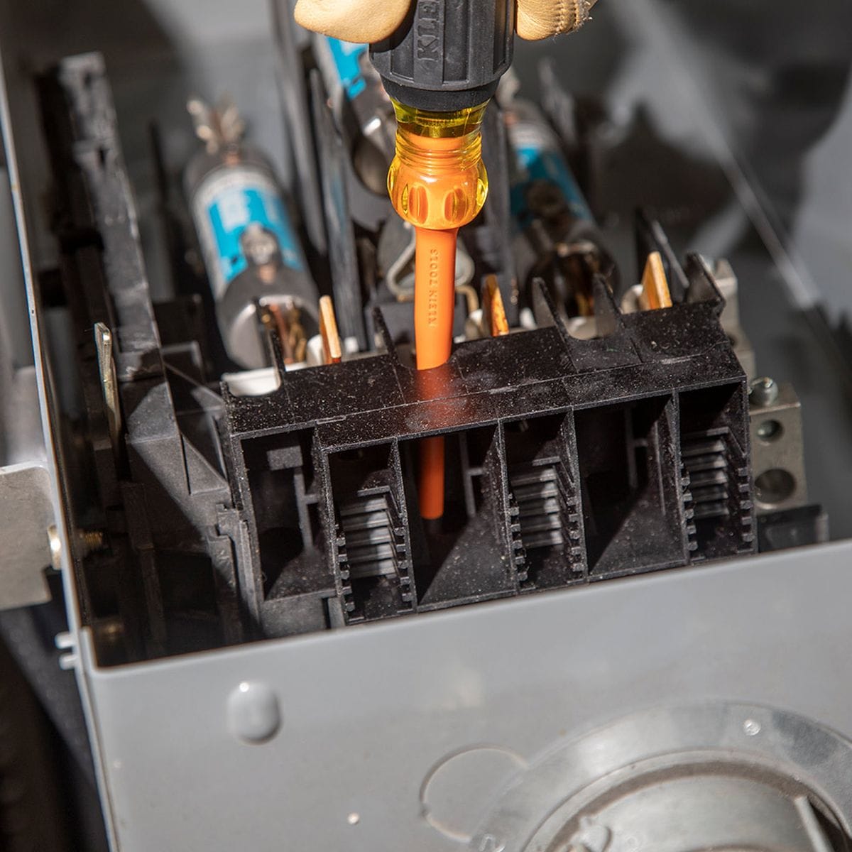 An electrician wearing tan leather safety gloves utilizes a Klein Tools 1000V rated orange slim-tip insulated screwdriver to drive a terminal screw deep within a compact industrial disconnect block, illustrating the flush sheathing profile's capacity to access tight electrical junctions without binding.