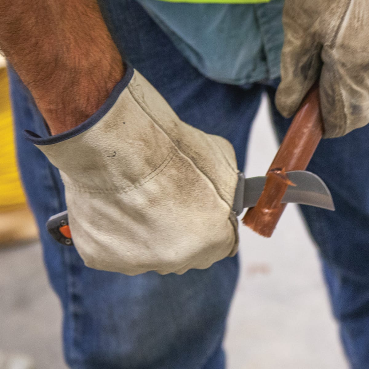 An action lifestyle shot of a professional electrician wearing heavy-duty work gloves using the Klein 44218 utility knife with a 44219 replacement blade. The worker is shown stripping thick copper cable, demonstrating the blade's high-performance cutting capability and ergonomic control for industrial skinning tasks.