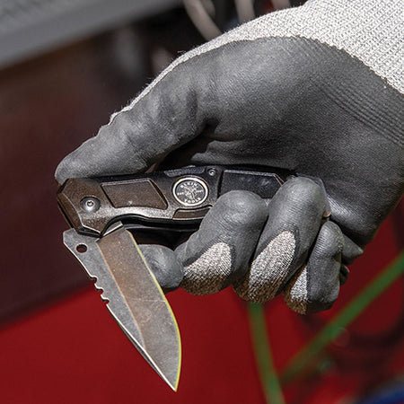 A worker wearing a textured gray polyurethane-coated grip glove grips the black aluminum handle of the folding pocket knife while deploying the stainless steel drop point blade, illustrating smooth bearing-assisted flipping mechanics and jobsite hand safety protocols.