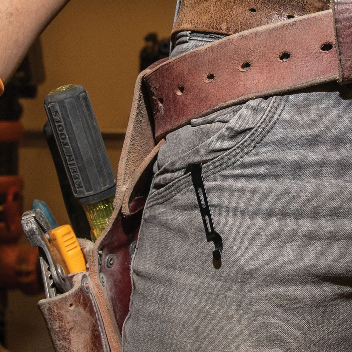 A field view of a tradesperson working on a site layout, highlighting the low-carry black pocket clip of the folding electrician knife tucked deeply into the front pocket of their gray canvas work pants next to a leather tool pouch stocked with screwdrivers and adjustable pliers.