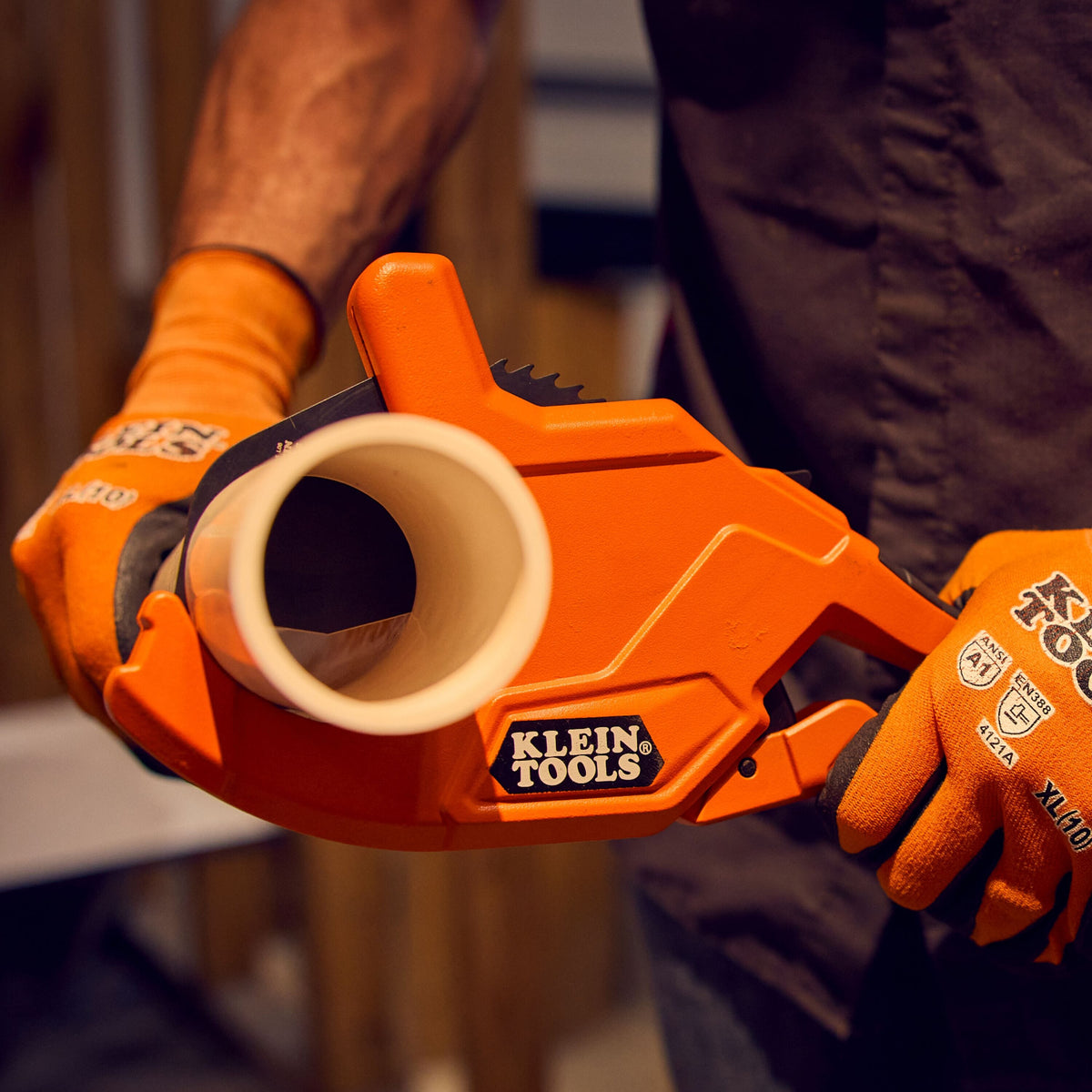 A real-world jobsite action shot showing a professional tradesperson wearing branded orange canvas utility safety gloves holding a white Schedule 40 PVC plastic conduit tube inside the deep structural jaw of the orange Klein Tools ratcheting cutter tool to prepare a plumb square line cut.