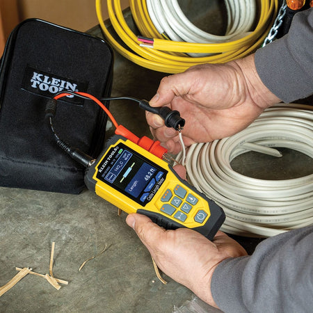 Action shot of the Klein Tools cable length meter being used to measure a coil of white 14/2 Romex electrical wire. The technician is shown using the heavy-duty alligator clips to connect to the copper conductors, with the meter displaying a real-time length measurement of 48.2 feet.