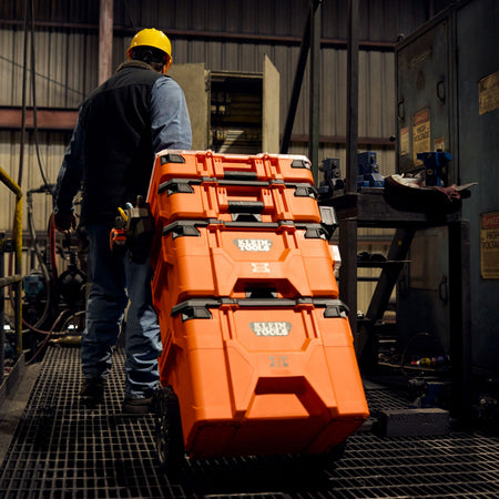 Action shot of a professional worker in a yellow hard hat rolling a fully stacked Klein Tools MODbox modular storage system across an industrial metal grate floor.
