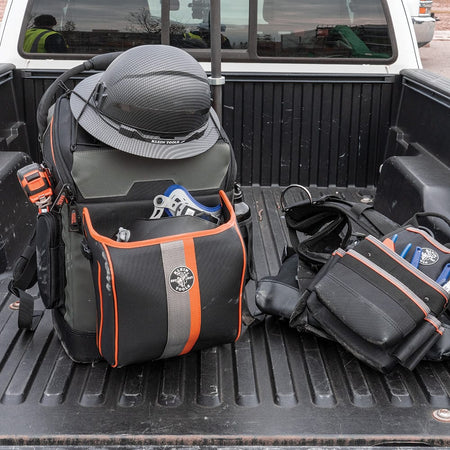 A lifestyle shot of the Klein Tools backpack resting in the bed of a pickup truck alongside a tool belt. The image shows the backpack's removable front pocket holding a welding helmet, highlighting the bag’s versatility for transporting trade-specific gear.