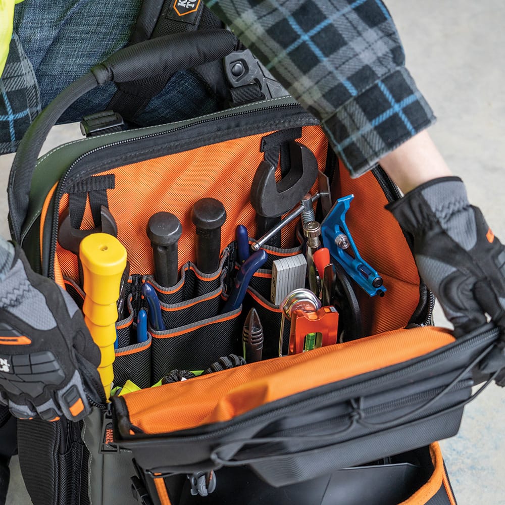 A close-up view of a technician reaching into the main compartment of the tradesman backpack. The high-visibility orange interior is shown fully organized with various hand tools, including wrenches and levels, emphasizing the 27 tiered pockets for superior tool accessibility.