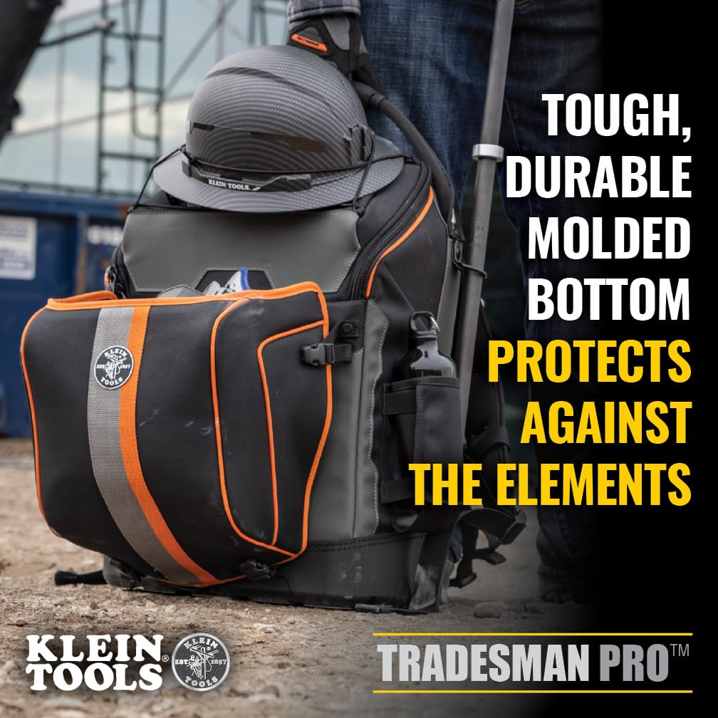 An action lifestyle shot of the Klein Tools tool bag on a construction site. The image demonstrates the backpack carrying a hard hat on top and a spud wrench on the side, highlighting the tough molded bottom that protects gear from dirt and debris on the ground.