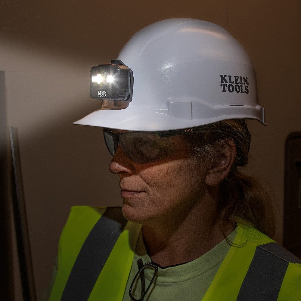 An action shot of a professional wearing a white Klein hard hat with the headlamp attached to the front bracket, illuminating a dark workspace.