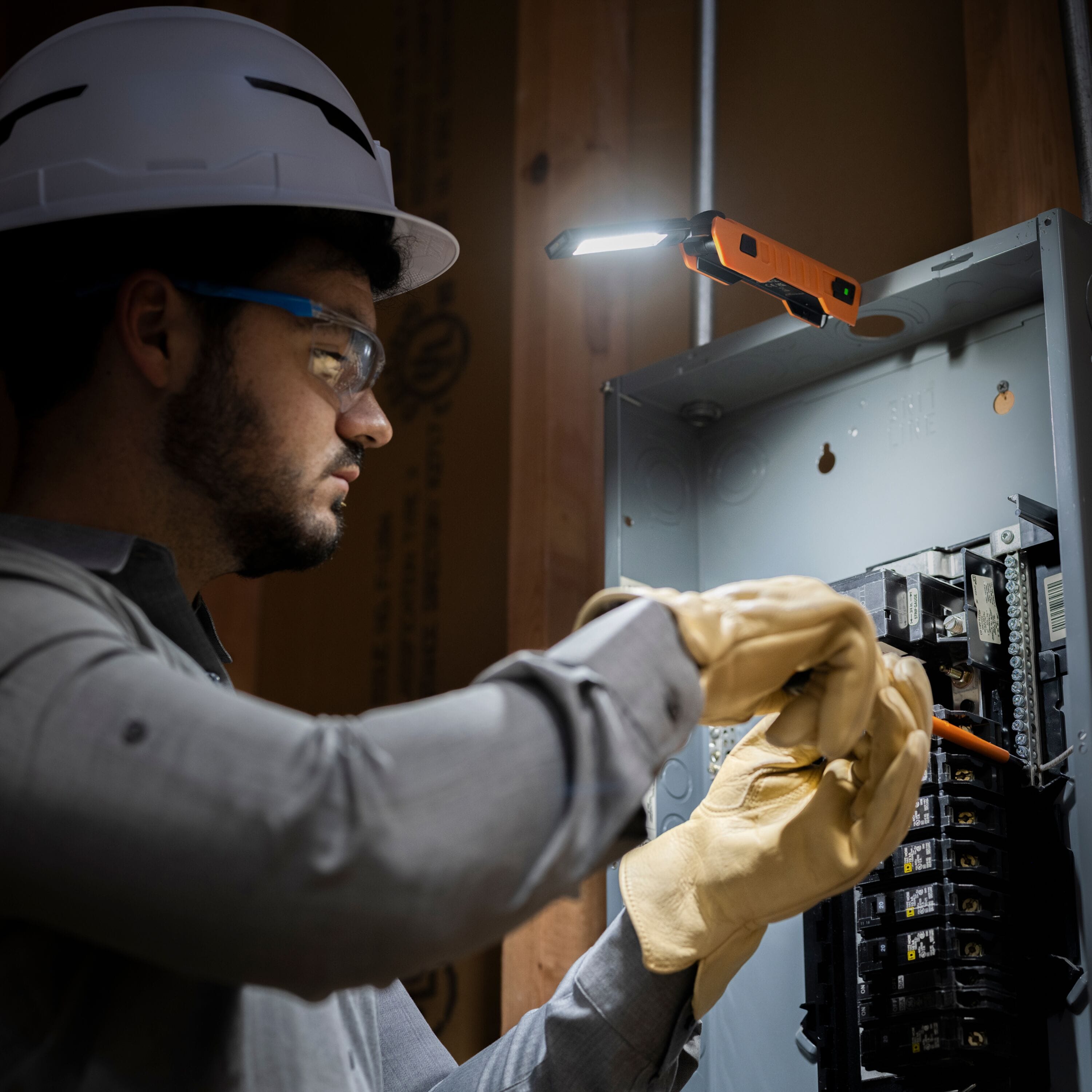 An operational demonstration of the Klein Tools rechargeable work light magnetically mounted to an open metal electrical panel, providing hands-free illumination for an electrician wearing a hard hat and safety gear.