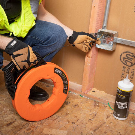 A worker kneeling on the floor feeding the fish tape into a low wall electrical box with a bottle of pulling lubricant nearby.