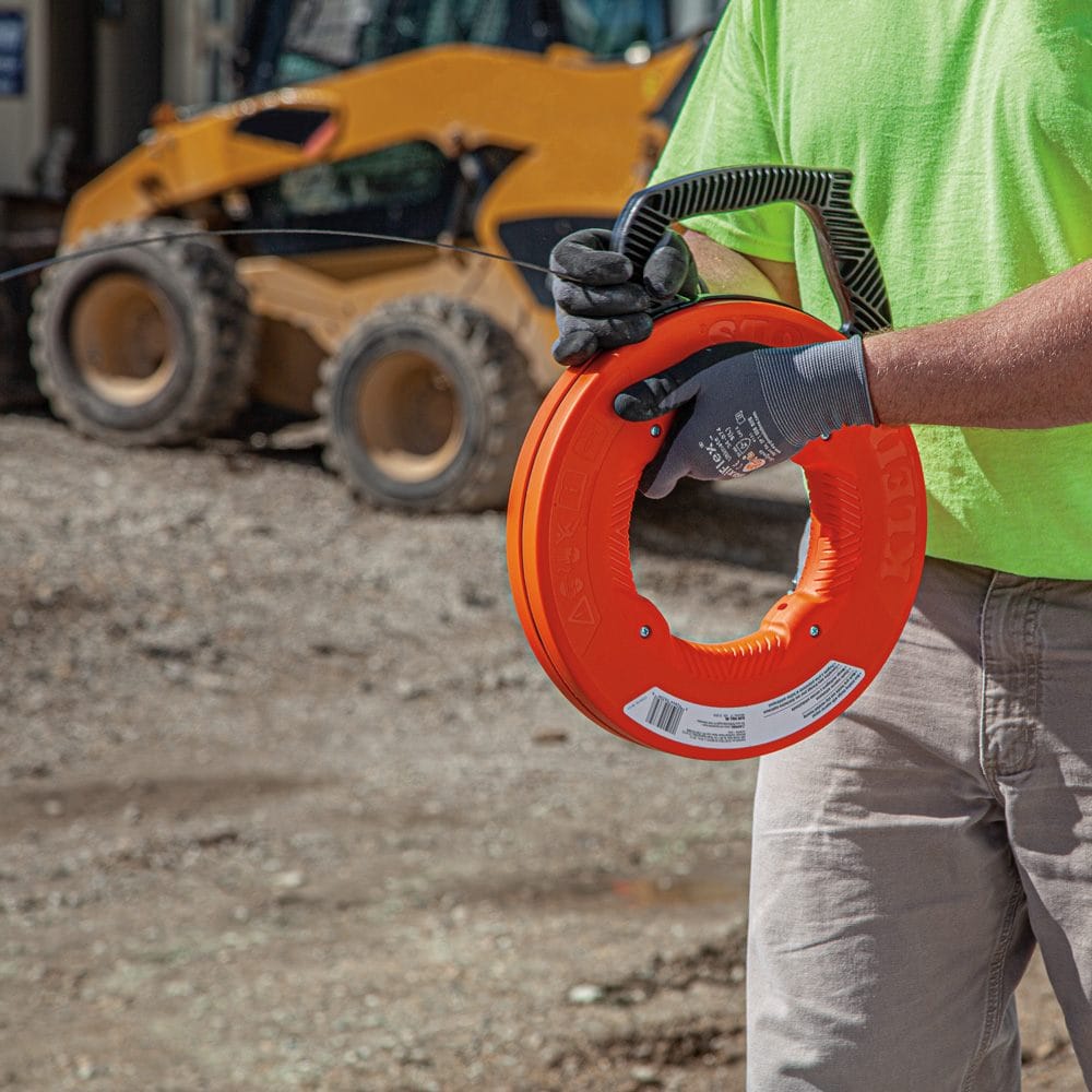 A worker wearing gloves holding the orange fish tape reel on an outdoor construction site with heavy machinery in the background.
