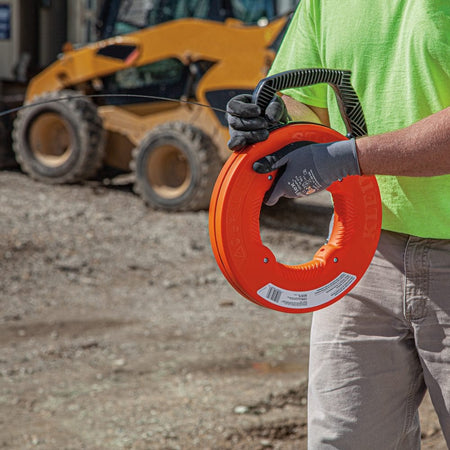 A worker wearing gloves holding the orange fish tape reel on an outdoor construction site with heavy machinery in the background.