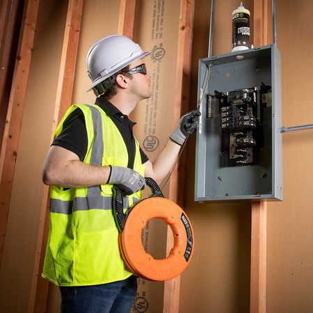 A technician wearing a hard hat and safety vest feeding the steel fish tape into an open electrical panel mounted on wooden studs.