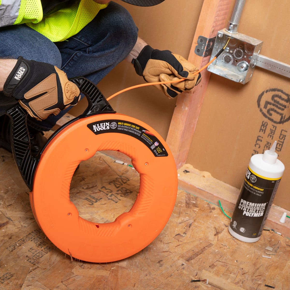An electrician using the Klein Tools fiberglass fish tape to pull wire through a junction box on a construction site, shown alongside Klein Tools premium synthetic polymer wire pulling lubricant for maximum efficiency.