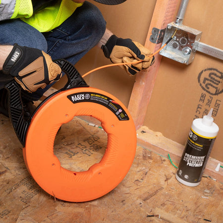 An electrician using the Klein Tools fiberglass fish tape to pull wire through a junction box on a construction site, shown alongside Klein Tools premium synthetic polymer wire pulling lubricant for maximum efficiency.