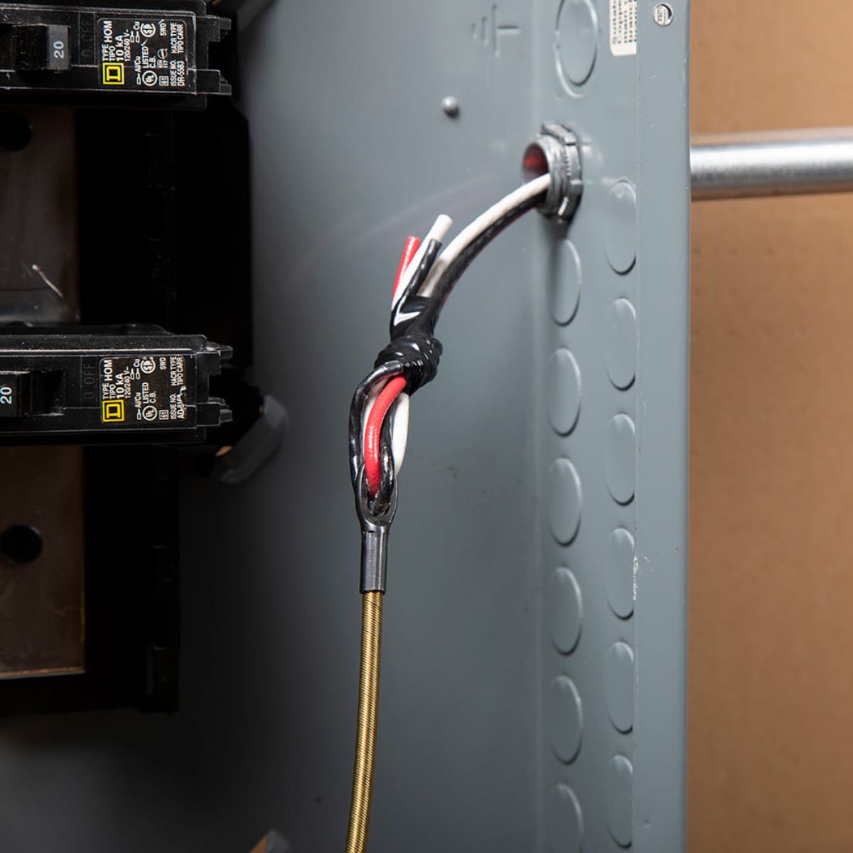 A close-up view of a spiral steel leader on a Klein Tools fish tape being used to pull electrical wires through a conduit knockout in a breaker panel, showing the secure attachment with electrical tape.