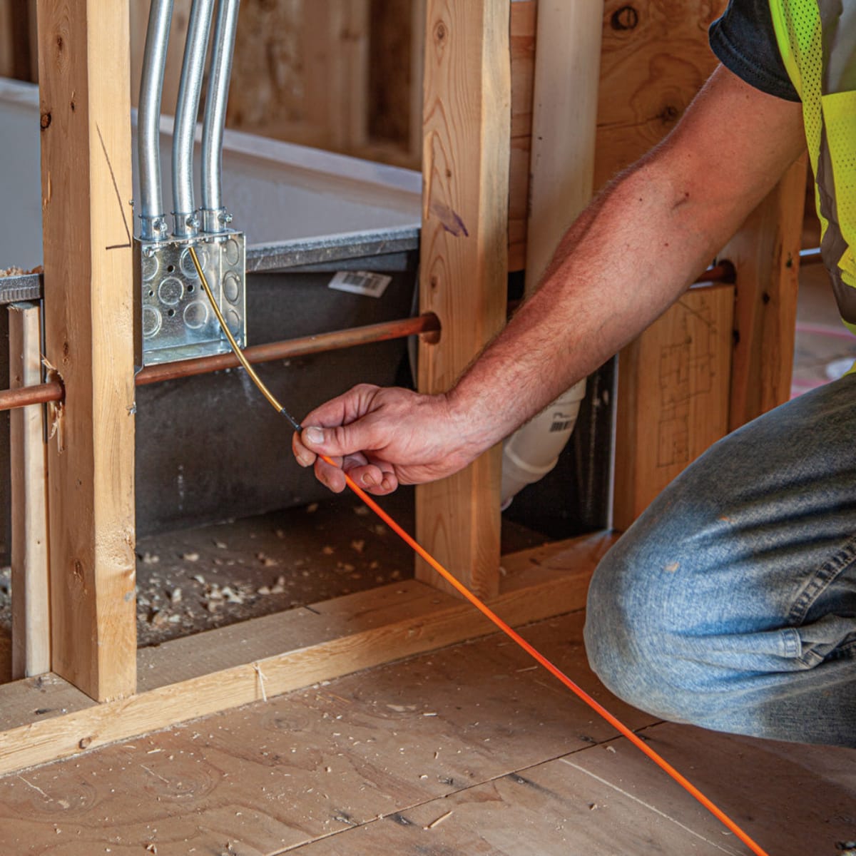 An electrician on a jobsite feeding the orange fiberglass fish tape into a metal electrical box mounted on wooden wall studs, illustrating the tool's flexibility and ease of use for residential wiring.