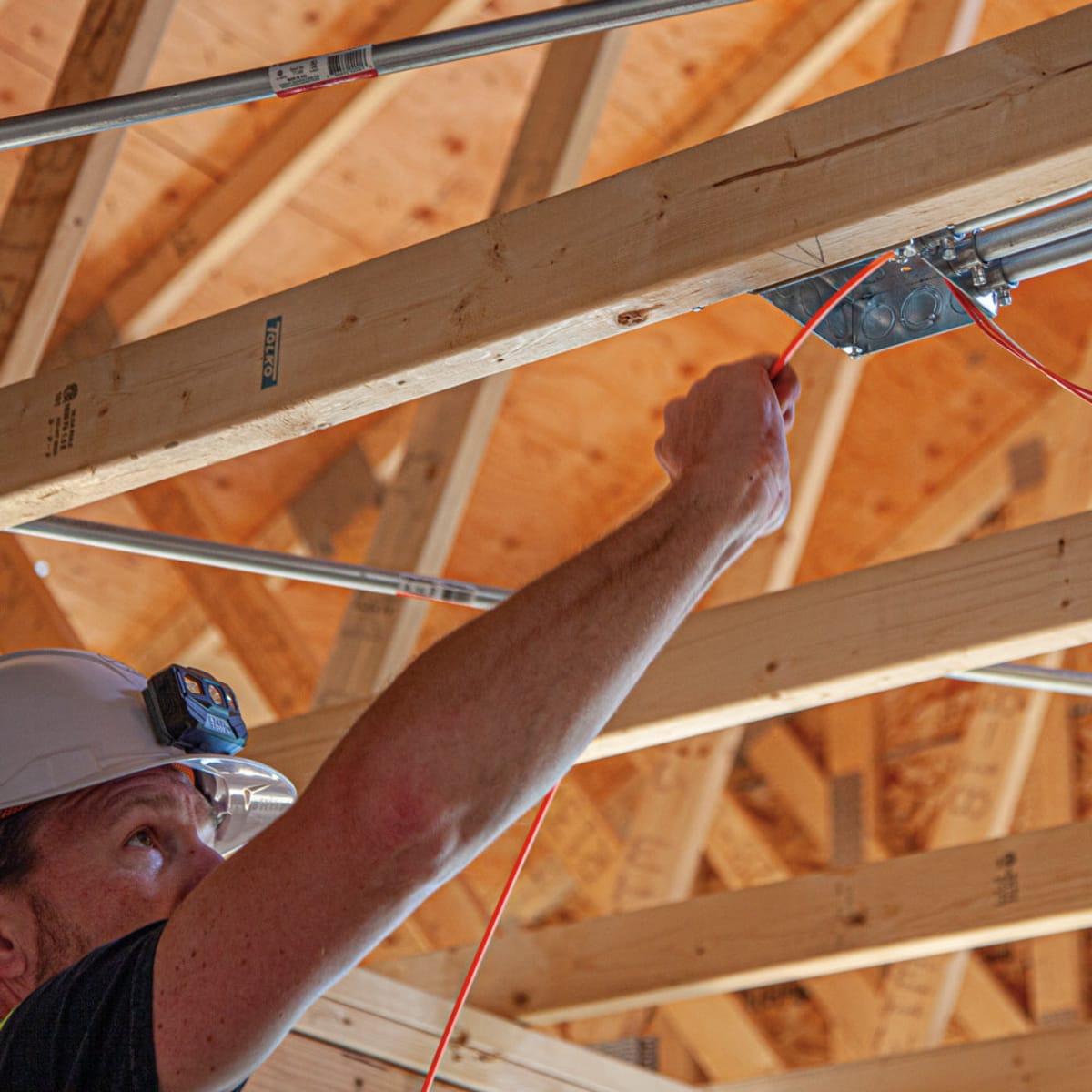 A professional worker in a hard hat navigating the Klein Tools fiberglass fish tape through conduit installed along wooden ceiling rafters, showcasing its high pushing power for overhead wire runs.