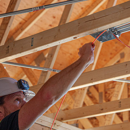 A professional worker in a hard hat navigating the Klein Tools fiberglass fish tape through conduit installed along wooden ceiling rafters, showcasing its high pushing power for overhead wire runs.