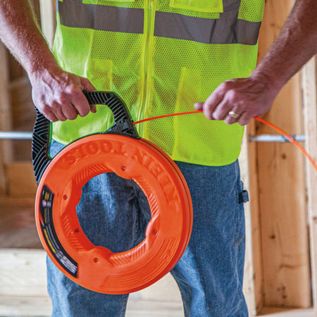 A front view of a worker holding the Klein Tools 100-foot fish tape case, demonstrating the ergonomic multi-position handle and the smooth payout of the orange multi-groove fiberglass tape.