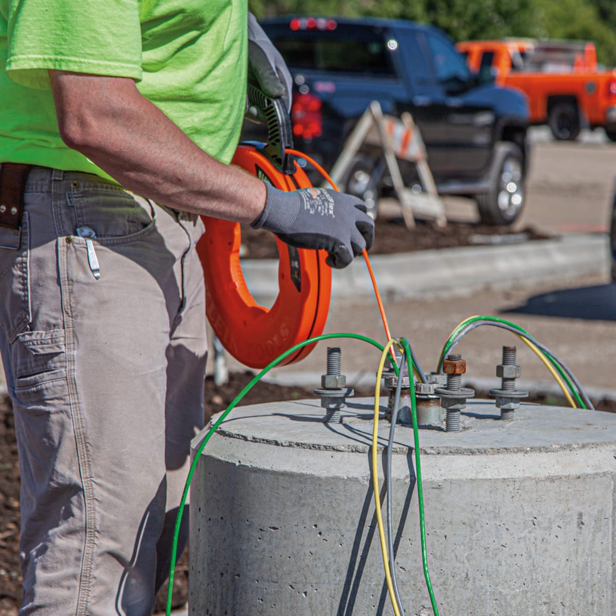 An outdoor application shot showing a contractor using the Klein Tools fiberglass fish tape to pull heavy-duty underground electrical cables through a concrete light pole base on a commercial jobsite.